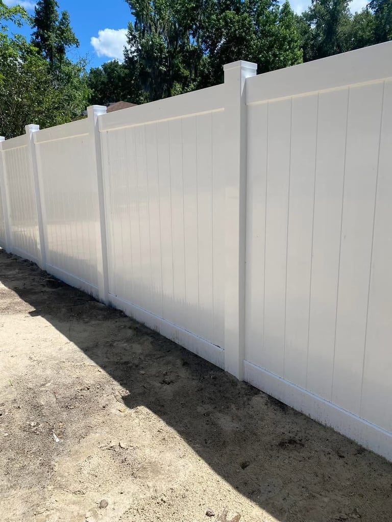 White vinyl privacy fence with vertical panels installed along dirt ground with trees and blue sky in background