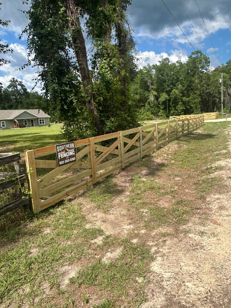 Wooden ranch fence with Southern Fencing sign along a rural driveway with trees and a house in the background