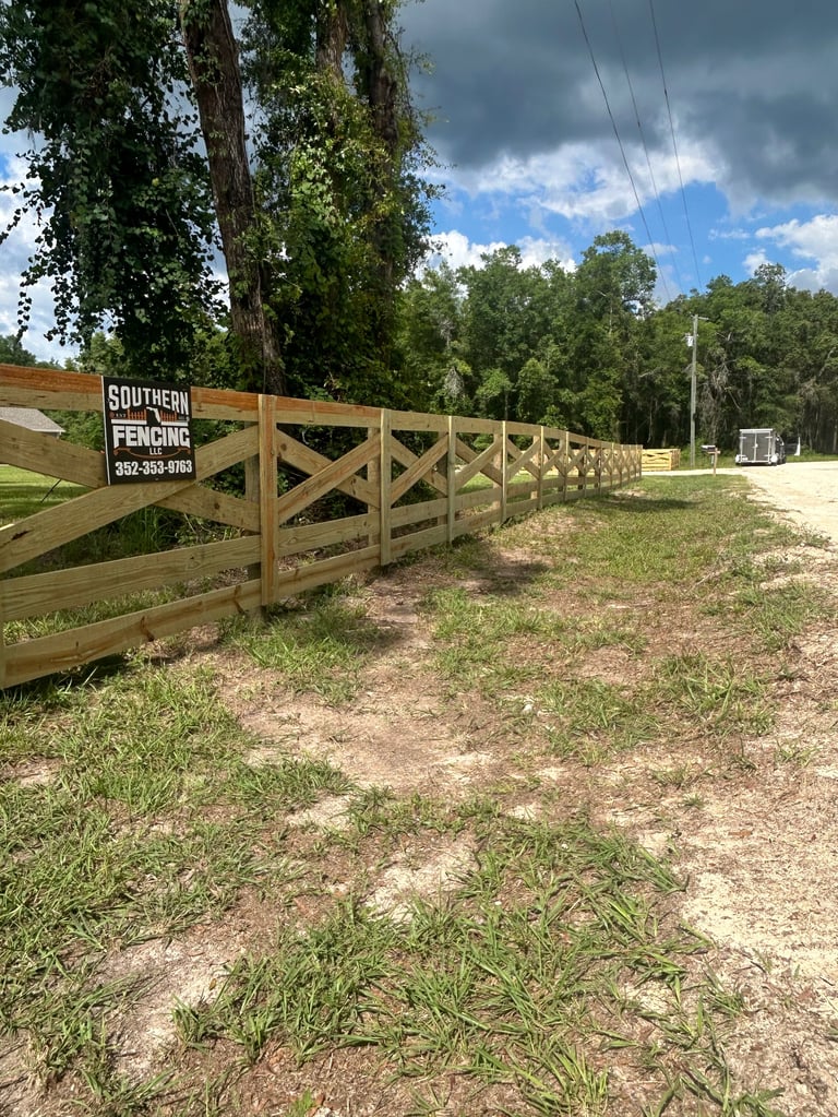 Wooden ranch fence with X-pattern design along a rural property under blue sky with clouds and tall trees