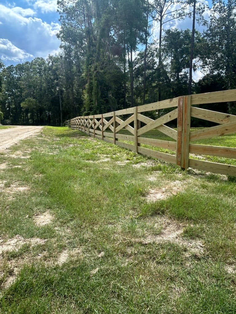 Long wooden ranch fence with X-pattern rails stretching through a grassy field lined with tall trees under a blue sky